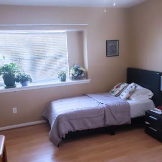 A cozy and well-lit bedroom featuring a bed, plants by the window, and simple furniture.