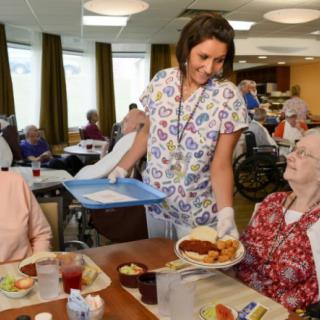 A caregiver serves a meal to residents in a nursing home dining area.