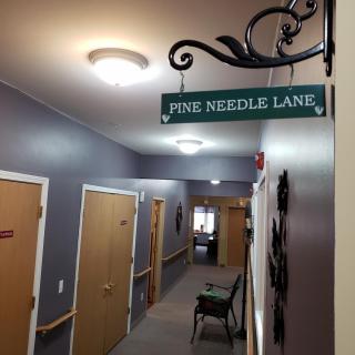 An indoor hallway featuring a sign that reads 'Pine Needle Lane' with several doors and overhead lighting.