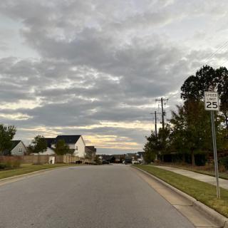 A quiet suburban street under a cloudy sky, with houses lining the road and trees on the side.