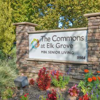 Sign for The Commons at Elk Grove, a senior living community, surrounded by plants and flowers.