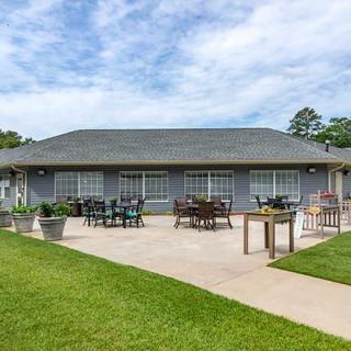 A building with a patio area featuring tables and chairs, surrounded by a well-maintained lawn under a partly cloudy sky.