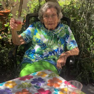 An elderly woman is sitting outdoors in a garden, raising a glass in a cheerful toast while wearing colorful clothing.