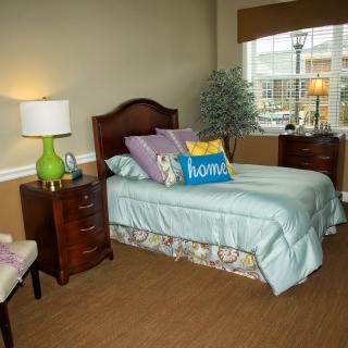 A cozy bedroom featuring a single bed with decorative pillows, a nightstand with a green lamp, and a window offering natural light.