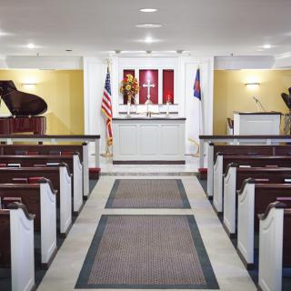 A small chapel interior with pews facing an altar, flanked by pianos and flags.