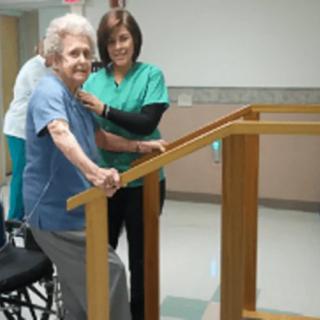 An elderly woman receives assistance from a caregiver during physical therapy exercises.