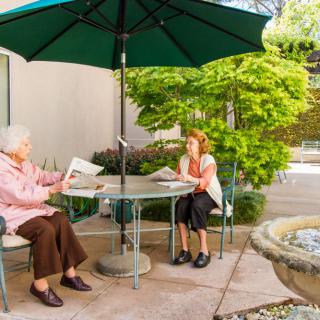 Two people sitting at a patio table under an umbrella, reading newspapers in a garden setting.