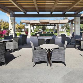 An outdoor patio area featuring wicker tables and chairs, set under a shaded pergola with a garden in the background.