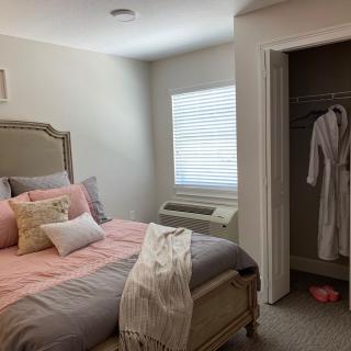A cozy bedroom with a neatly made bed, featuring pink and gray tones, alongside an open closet displaying a hanging robe.