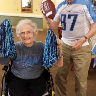 An elderly person in a wheelchair holds blue pom poms while another person stands nearby holding a football.