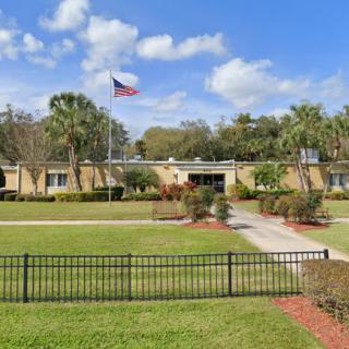 A single-story building with an American flag on its roof, surrounded by a well-maintained lawn, palm trees, and a black fence under a partly cloudy sky.