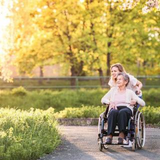 A caregiver assists an elderly person in a wheelchair during a sunny day in the park.