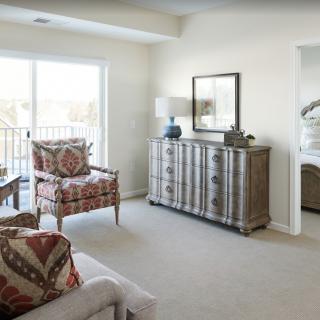 A cozy living room featuring patterned armchairs, a rustic sideboard, and large windows letting in natural light.