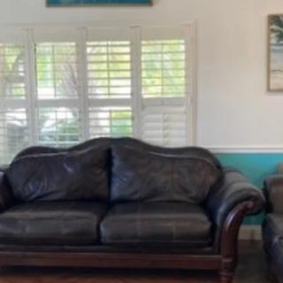 A cozy living room featuring a dark leather sofa and armchair, complemented by wall art and shuttered windows.