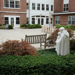 A serene courtyard featuring a statue and a bench, surrounded by greenery and brick architecture.
