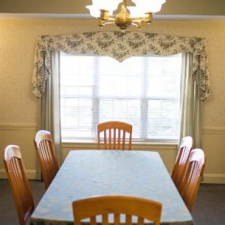 A dining room with a wooden table and six chairs, featuring a large window with patterned curtains and a chandelier overhead.