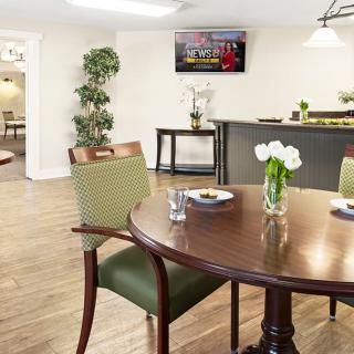 A cozy dining area with a wooden table, green chairs, and a flower vase centerpiece, set against a background with a television and elegant decor.