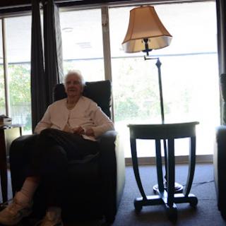 An elderly person sitting comfortably in an armchair beside a lamp in a well-lit indoor space.
