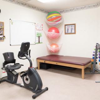 A therapy room equipped with exercise equipment, including a stationary bike, therapy balls, weights, and a treatment table.