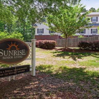 A sunny day view of a building, with a 'Sunrise Senior Living' sign in the foreground, surrounded by trees and well-maintained landscaping.