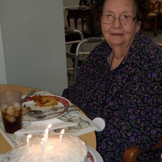 An elderly woman sitting at a dining table with a cake and lit candles, celebrating her birthday.
