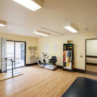 A well-lit rehabilitation room featuring exercise equipment and parallel bars on a wooden floor.