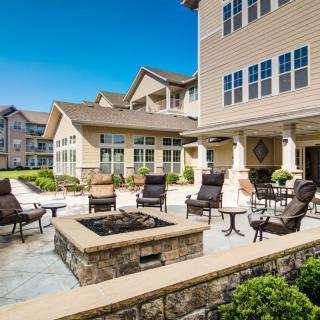 A well-designed residential patio area with a fire pit surrounded by chairs, set against the backdrop of multi-story buildings and clear blue sky.