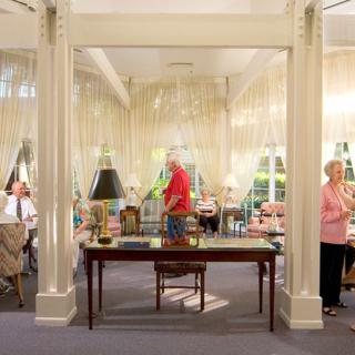A well-decorated interior space featuring people, a central desk with a lamp, and large windows.