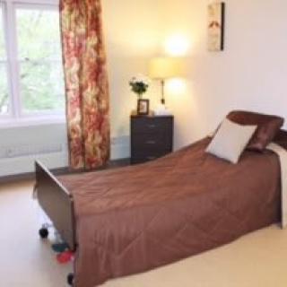 A cozy bedroom featuring a single bed with a brown cover, a nightstand with a lamp, and floral curtains by the window.