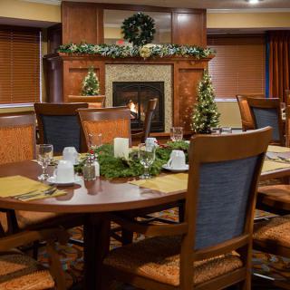 A cozy dining room with a round table set for a meal, decorated with holiday greenery and candles near a fireplace.