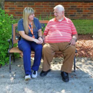 A caregiver and an elderly man sitting on a bench outdoors having a conversation.