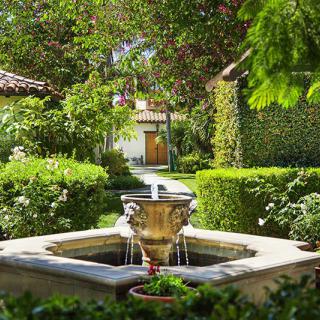 A tranquil garden scene featuring a central fountain surrounded by lush greenery and a pathway leading to a charming building.