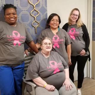A group of women wearing matching T-shirts with a pink ribbon design, posing indoors to support breast cancer awareness.