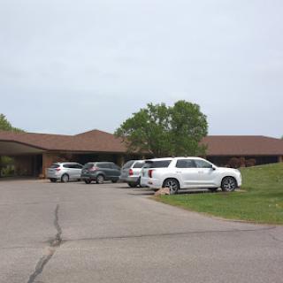 A view of a building with several parked cars in its lot, surrounded by greenery.
