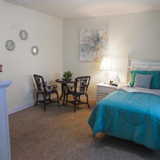 A cozy bedroom with a turquoise bedspread, a small table and two chairs, a lamp, and wall decor.