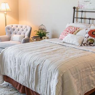 A cozy bedroom with a neatly made bed, an upholstered chair, and a standing lamp.
