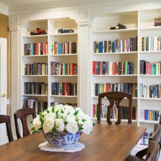 A cozy library room with white bookshelves filled with books and a wooden table adorned with a vase of flowers.
