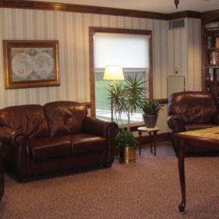 A cozy living room featuring leather sofas and armchairs, a central lamp, potted plants, and a bookshelf in the corner.