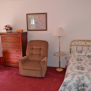 A vintage-styled bedroom with a brown armchair, a chest of drawers, and a bed with floral bedding.