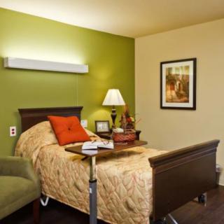 A cozy nursing home room with a bed, a side table with a lamp, and an armchair.
