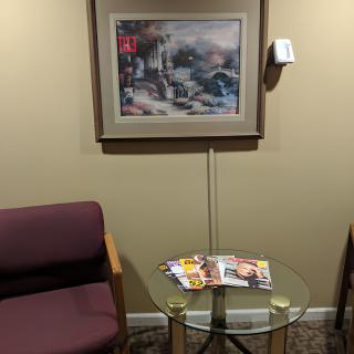 A waiting room with a chair, glass table holding magazines, and a painting on the wall.