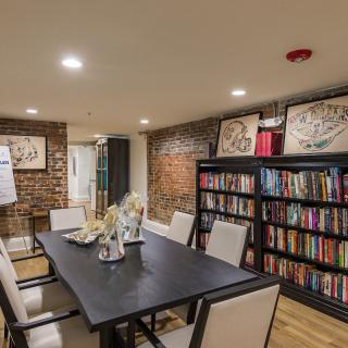 A cozy dining room with a large table, surrounded by chairs and decorated with bookshelves and artwork on exposed brick walls.