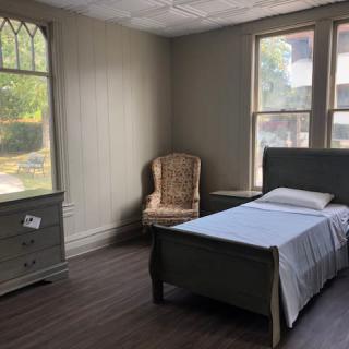 A minimalist bedroom with a simple bed, chair, dresser, and large windows.