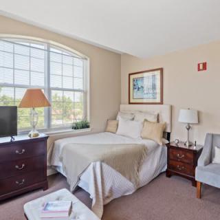 A cozy bedroom with a large window, featuring a bed, dresser, lamps, and seating area.