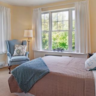 A cozy bedroom with a large window, featuring an armchair and a bedside lamp.