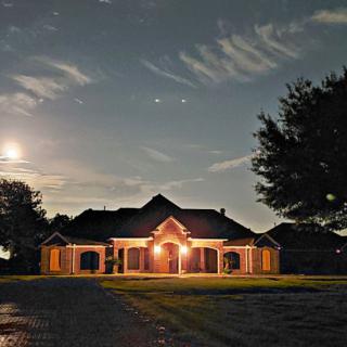 A house illuminated by outdoor lights under a night sky with clouds and the moon.