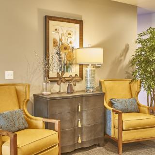 A cozy lounge area featuring two yellow chairs, a central cabinet with a lamp, and decorative elements such as a painting and indoor plant.