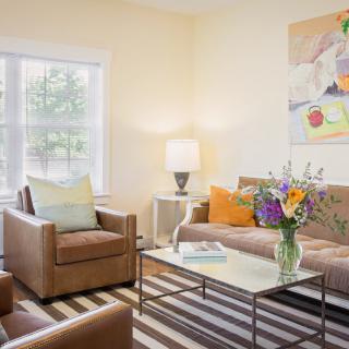 A cozy living room with modern furniture, including a sofa and two chairs, accented by colorful pillows and a bouquet of flowers on the coffee table.