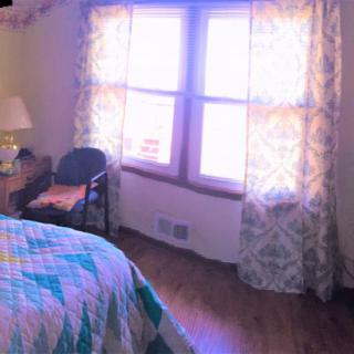 A cozy bedroom with a quilt-covered bed, a chair, and patterned curtains framing a sunlit window.