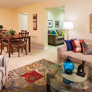 A cozy living room with a dining area, featuring a sofa and chairs, bright decor, and a glass coffee table.